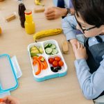 课后辅导 A young boy sits at a wooden table in school, enjoying a healthy lunch from a colorful lunchbox.